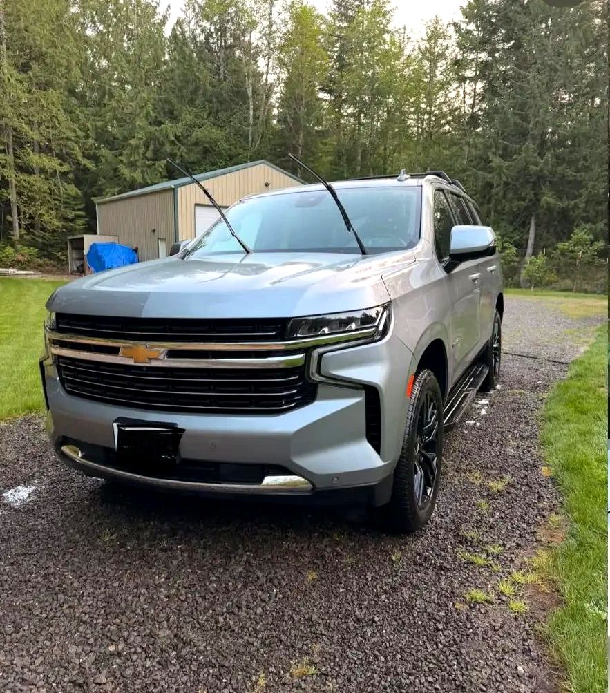 Freshly washed silver Chevy Tahoe parked on a gravel driveway among evergreen trees